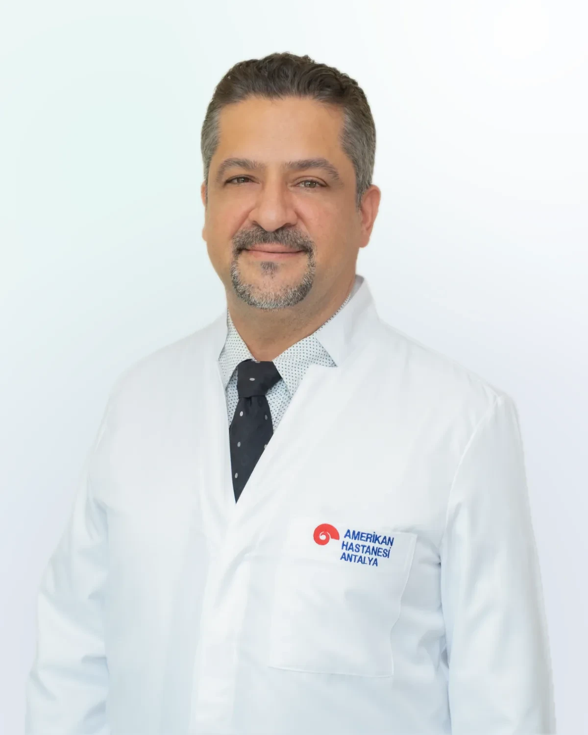Beyaz önlük ve kravat giymiş, göğsünde Amerikan Hastanesi Antalya logosu görünen erkek doktor portresi.